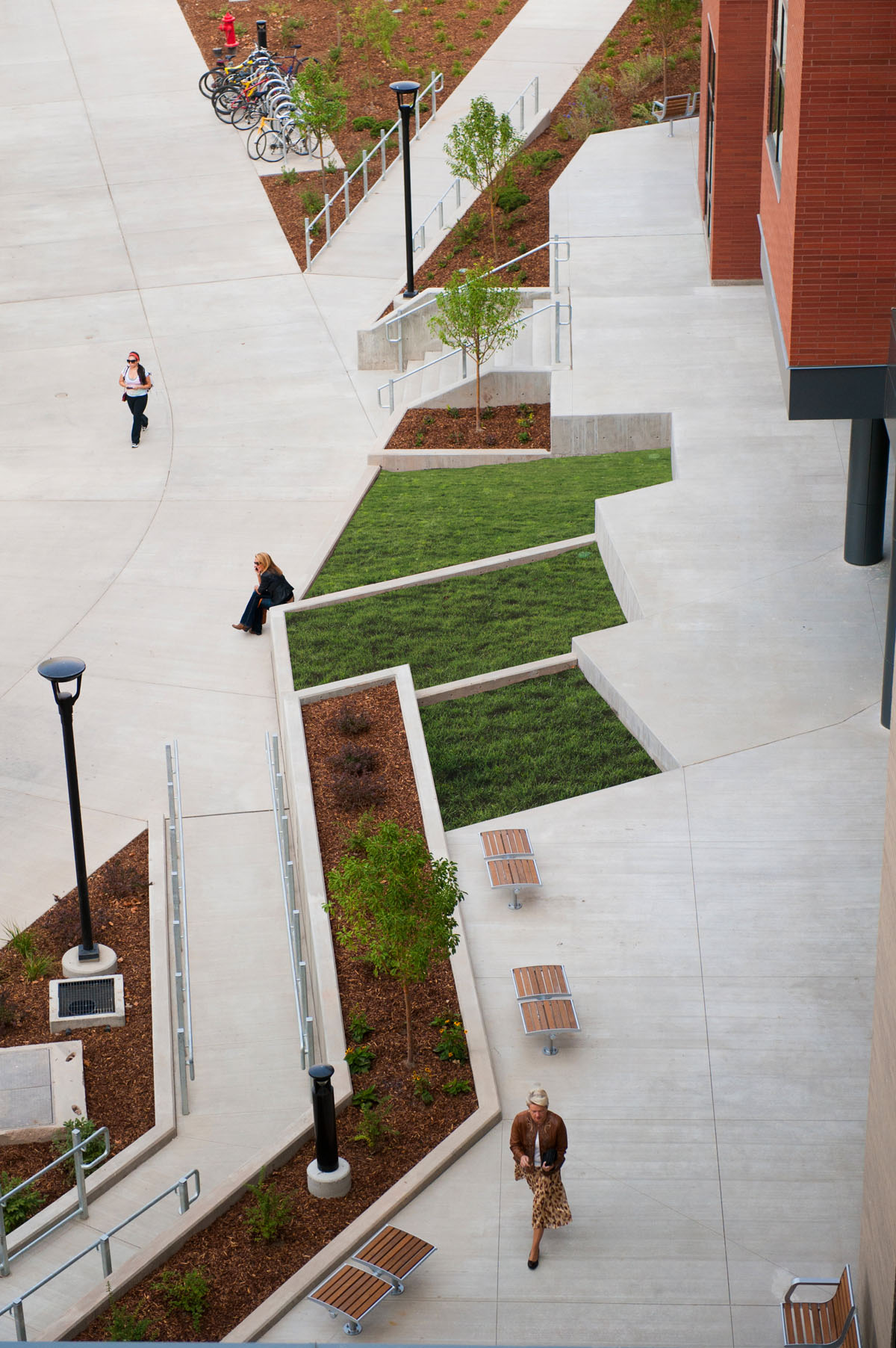 A woman walks next to the landscaping of the honors student housing building as another woman sits next to the grass and a student walks towards the building.