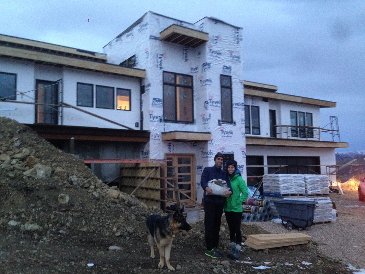 A German Shepherd, man, and woman stand in front of the modern home while it is under construction.