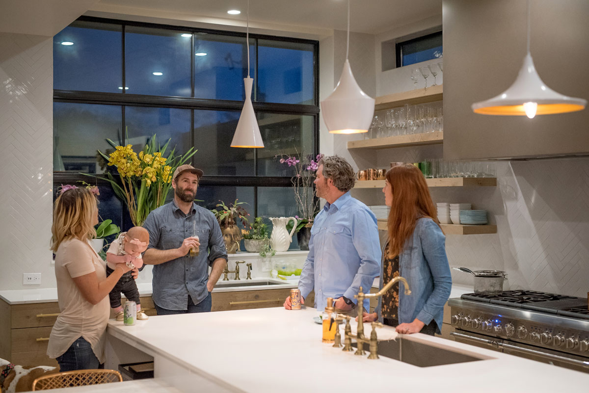 A woman and her baby, two men, and another woman stand around the kitchen island talking.